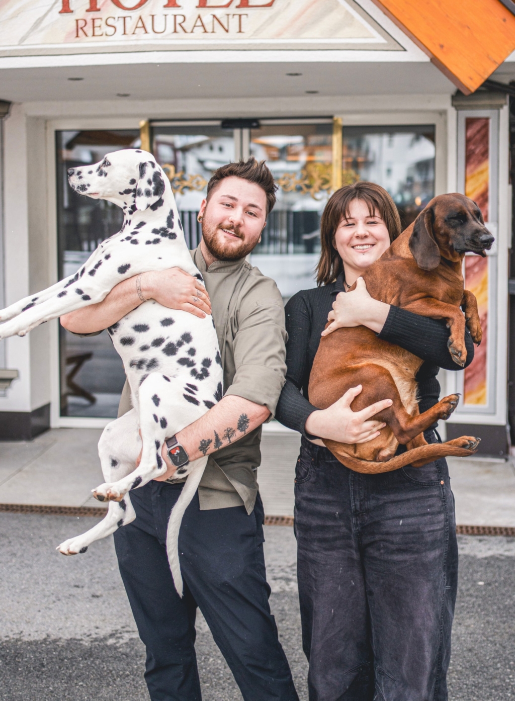 Hosts are standing in front of a restaurant holding two dogs. The man is carrying a Dalmatian and the woman a brown dog